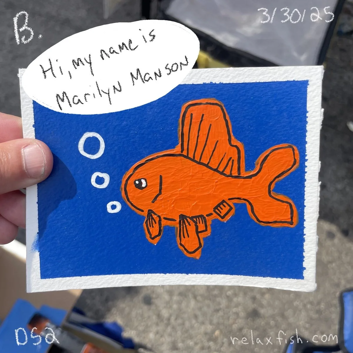 A hand holding a drawing of an orange fish with black outlines against a blue background. The fish has fins and a sad expression. There is a speech bubble saying, 'Hi, my name is Marilyn Manson.' The date 3/30/23 is written in the top right corner.