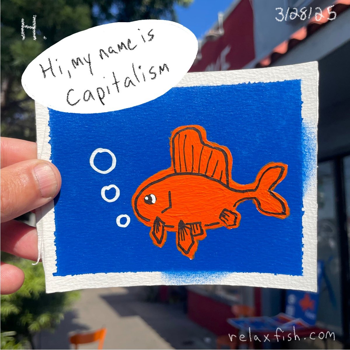 Drawing of a cartoon orange fish with fins, swimming in blue water with bubbles. Speech bubble says, 'Hi, my name is capitalism.' Hand holding the drawing outdoors, with a store and some trees in the background.