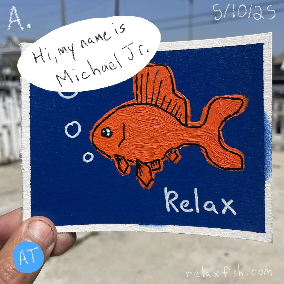 Handheld illustration of an orange fish with black outlines and bubbles, labeled 'relax,' with a speech bubble introducing itself as Michael J.R., against a dark blue background, at an outdoor location.