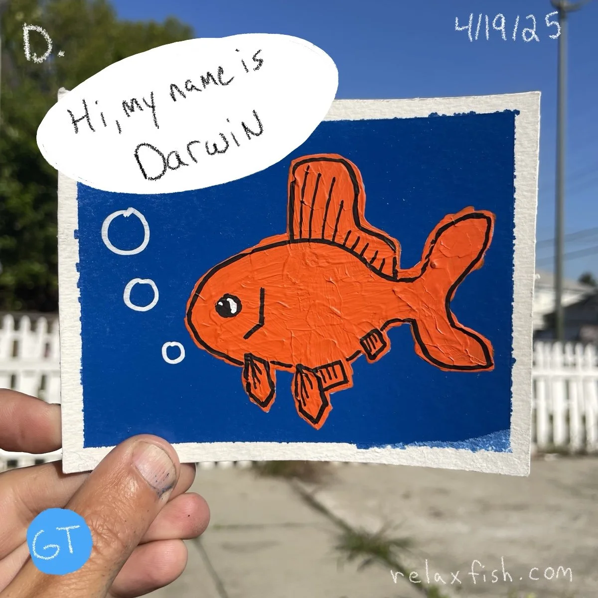 A colorful drawing of a cartoon orange fish with fins and bubbles, held up outdoors against a clear blue sky and white fence. Speech bubble says "Hi, my name is Darwin."