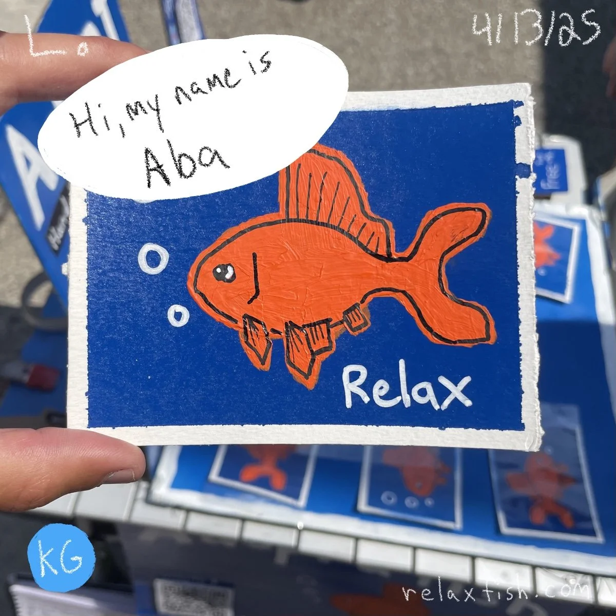 A colorful drawing of an orange fish with black outline, surrounded by bubbles, on a blue background. The drawing is on a square piece of paper with the word 'Relax' written at the bottom. A speech bubble on top says, 'Hi, my name is Aoba.' There are