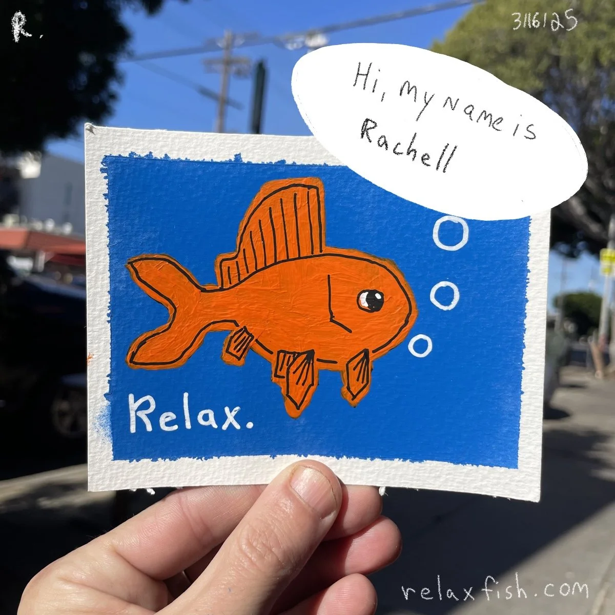 A colorful drawing of an orange fish with a blue background, bubbles, and the word "Relax" written beneath it. A speech bubble says, "Hi, my name is Rachell." Hand holding the artwork, with a street scene in the background.