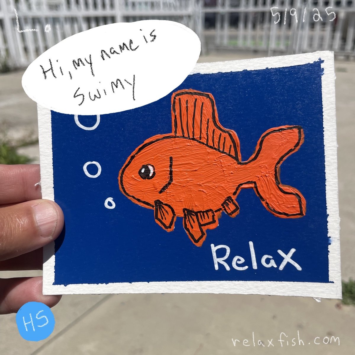 A handmade drawing of an orange fish with a smile, bubbles, and fins on a blue background, with the words "Relax" and a speech bubble saying "Hi, my name is Swimy". The drawing is on a square piece of paper held outdoors.