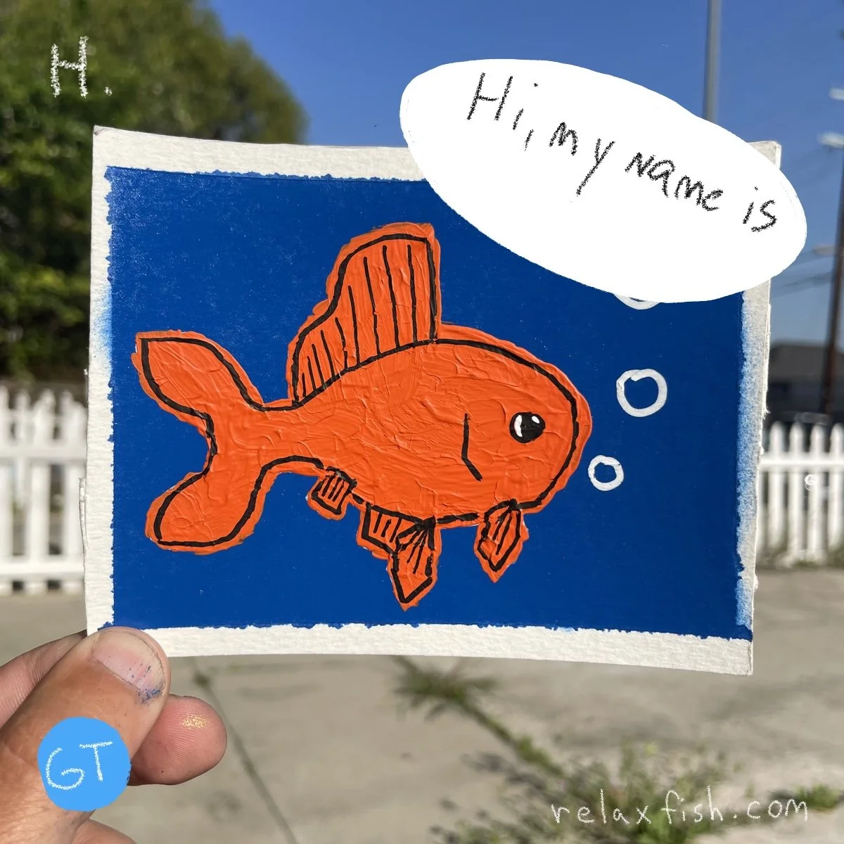 A hand holding a colorful drawing of an orange fish wearing a hat, with bubbles near its mouth, against a blue background. The fish has a speech bubble that reads, 'Hi, my name is'.