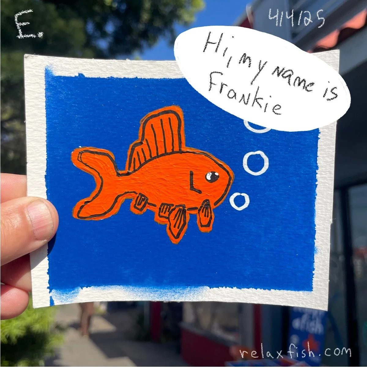 A colorful drawing of an orange fish with black outline and fins, depicted against a blue background. A speech bubble says, 'Hi, my name is Frankie.' The artist's hand is holding the drawing, and outdoor trees and buildings are visible in the backgro