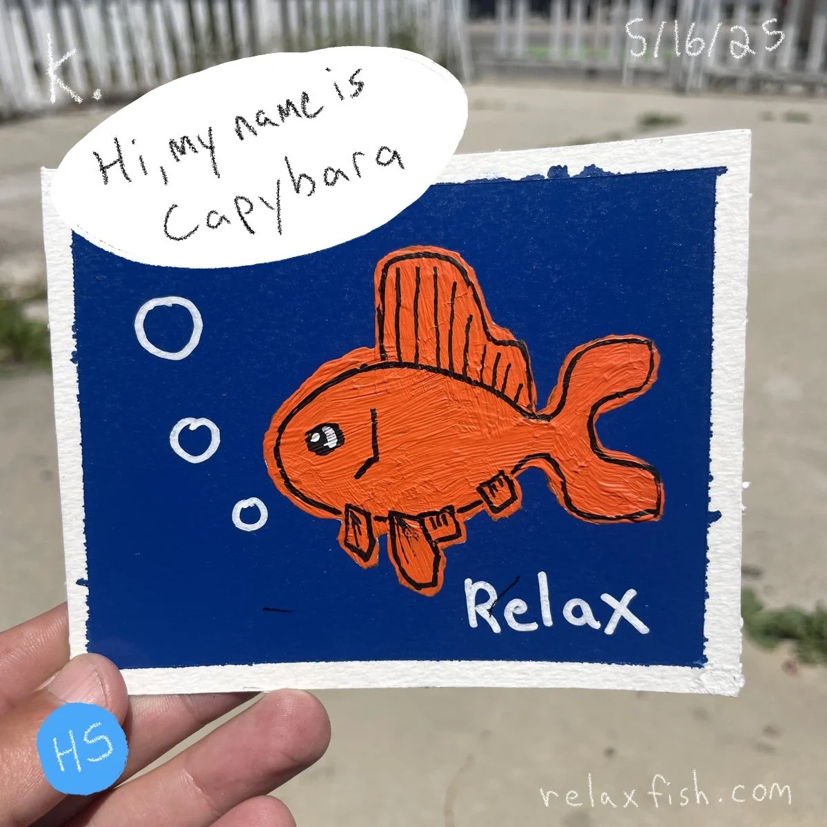 Colorful drawing of an orange fish with a dorsal fin, pectoral fins, and a tail, against a blue background. The fish is smiling, has a large eye, and the word 'Relax' written below. A speech bubble says, 'Hi, my name is capybara.' Hand holding the pa
