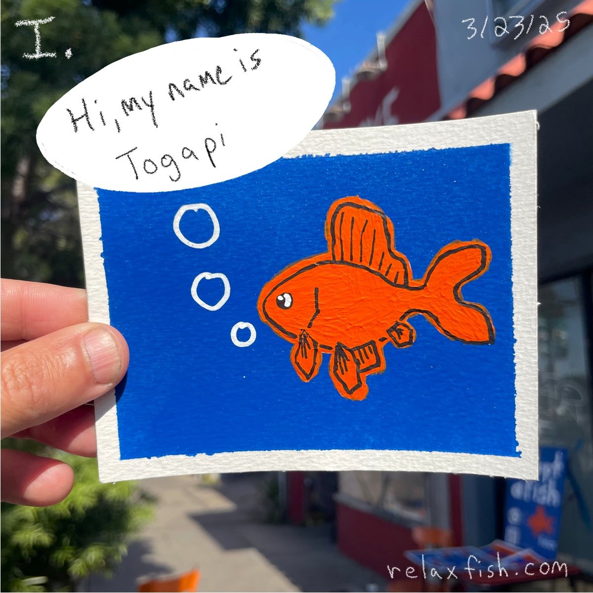 Hand holding a drawing of a goldfish in a fishbowl with a speech bubble that says, "Hi, my name is Togapi." The drawing is on a bright blue background, and there are some trees and a building in the background.
