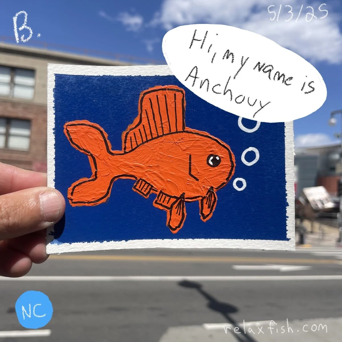 A hand holding a piece of paper with a colorful drawing of an orange fish wearing a top hat and a bow tie against a dark blue background. A speech bubble says, 'Hi, my name is Anchovy.' The background shows a city street with buildings, a lamp post, 