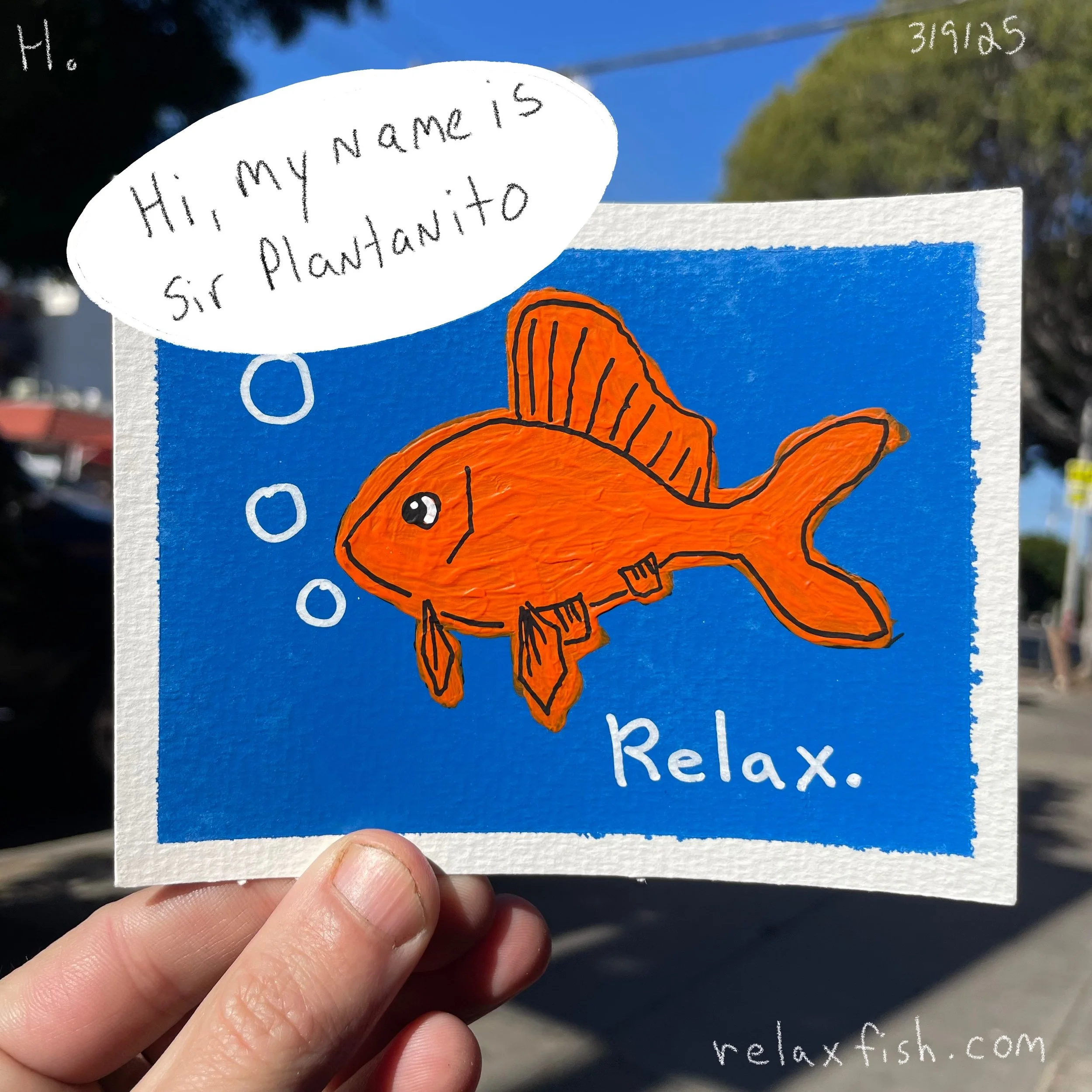 A colorful drawing of a fish with orange body and fins on a blue background. The fish has a speech bubble saying, "Hi, my name is Sir Plantanito." The word "Relax" is written below the fish. The drawing is held by a person's hand against an outdoor b