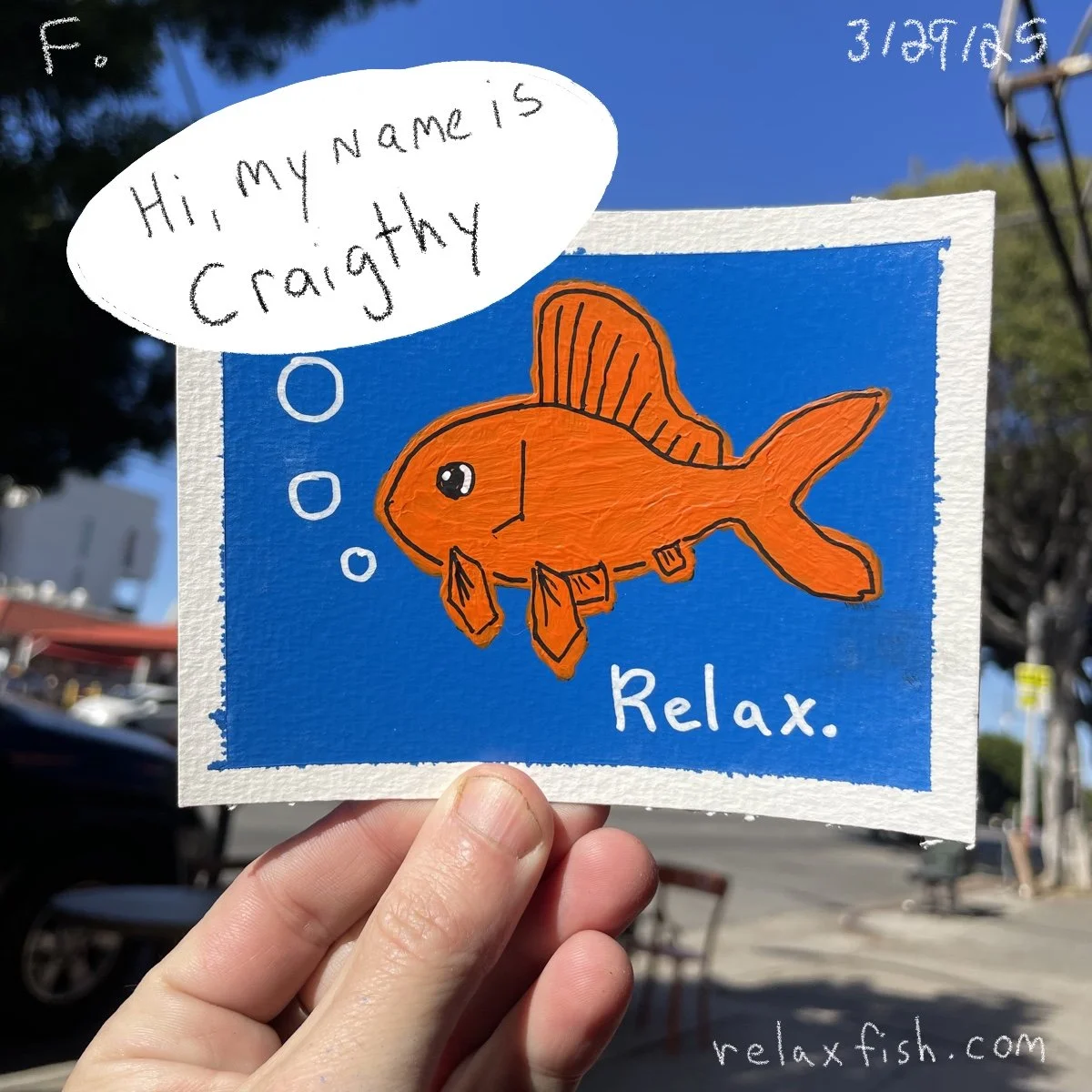 Hand holding a colorful illustration of a fish with a speech bubble that says, 'Hi, my name is Cragthy.' The drawing has a blue background with the word 'Relax.' written on it, and the website relaxfish.com is visible at the bottom.