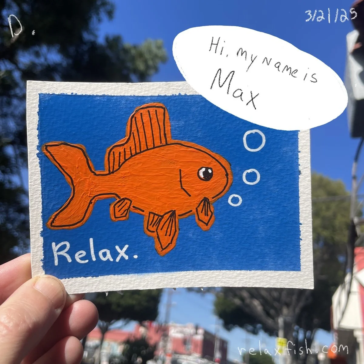 A hand holds a colorful drawing of an orange fish with a small hat, against a blue background. The drawing includes the text "Relax." and a white speech bubble that says "Hi, my name is Max." The date "3/2/23" is written in the top right corner.