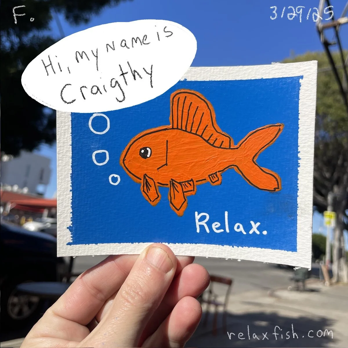 Hand holding a colorful drawing of a goldfish with the words "Relax" written on it. The fish appears to be thinking, with a speech bubble that says "Hi, my name is Crazyth". The background shows an outdoor setting with blue sky and trees.