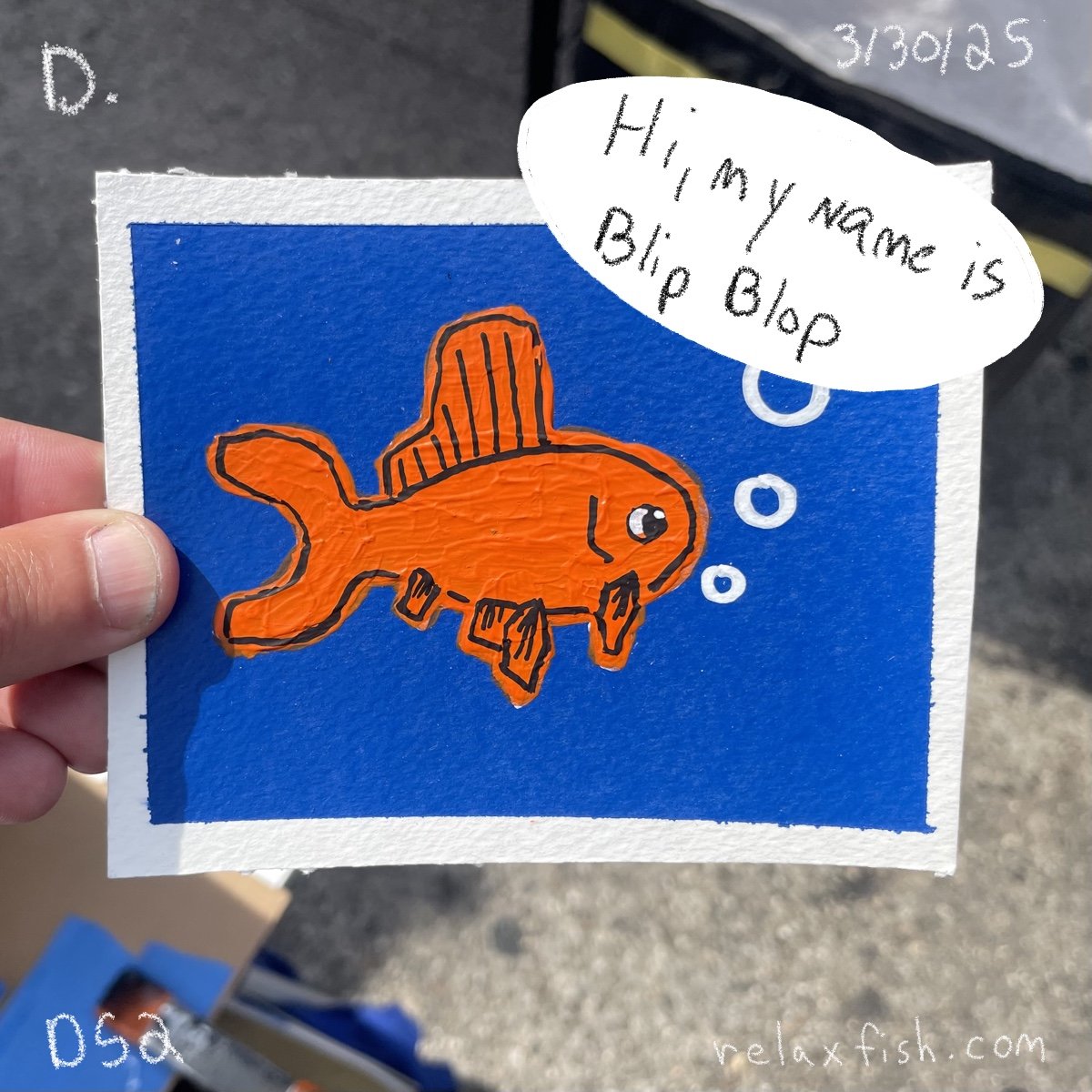A hand holding a blue card with a drawing of an orange fish with a hat, and a speech bubble saying 'Hi, my name is Bip Blob.' The background appears to be an outdoor setting with pavement. Handwriting on the top right says '3/30/25' and on top left '
