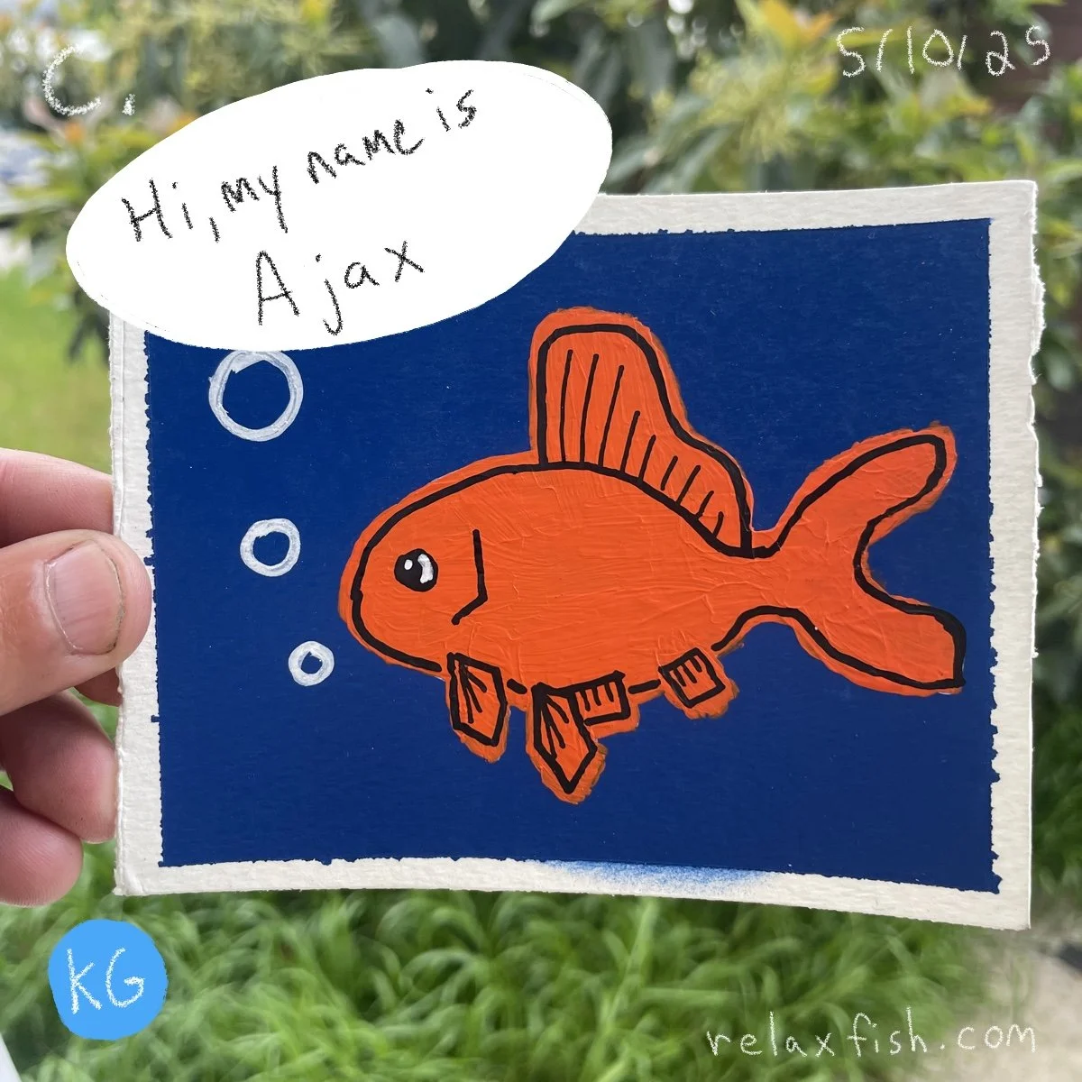 Children's drawing of an orange fish wearing a robotic suit, with a speech bubble saying, 'Hi, my name is Ajax.' The background is dark blue, and the drawing is on a piece of paper held by a person's hand outdoors. The paper has a blue sticker with '