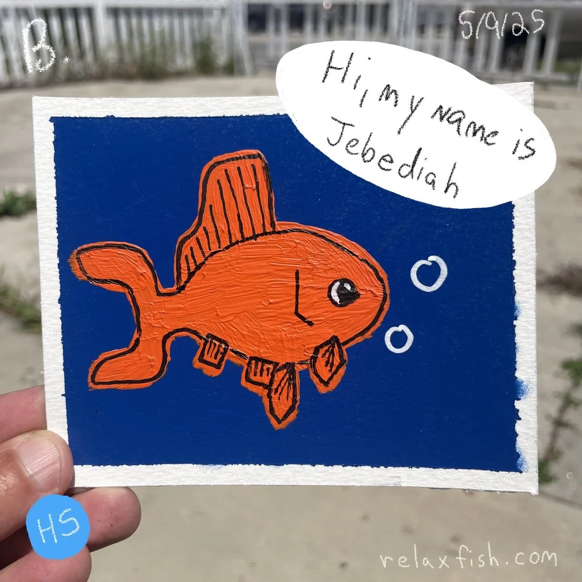 A colorful drawing of an orange fish wearing a pirate hat and a necklace, with bubbles around it, on a blue background. Handwritten text in a speech bubble says, "Hi, my name is Jebediah." The drawing is on a piece of paper held outdoors.