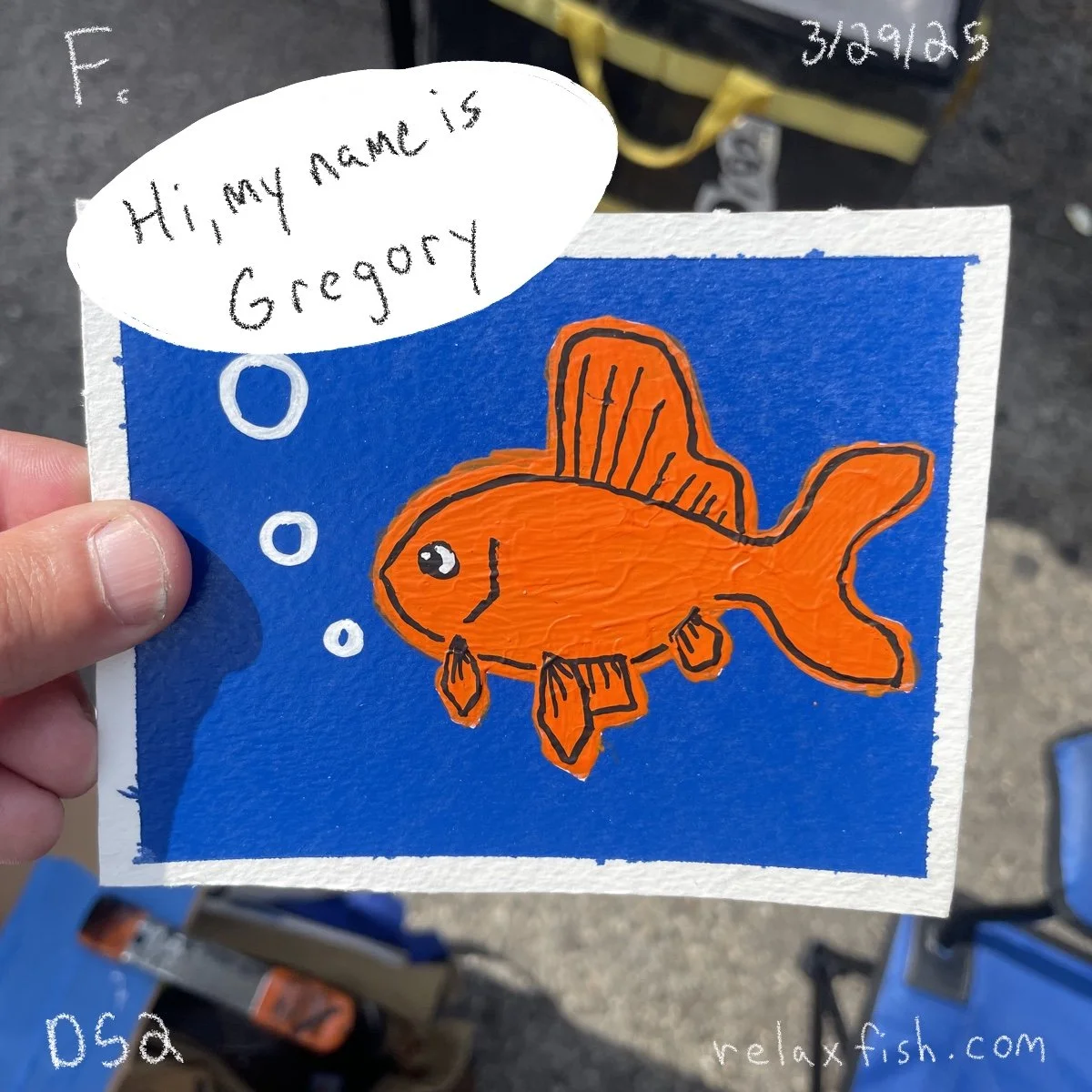 Colorful hand-drawn picture of an orange fish with black outlines on blue background, with a speech bubble saying "Hi, my name is Gregory." The drawing appears to be on a small square poster, held by a person's hand. The background shows a yellow obj