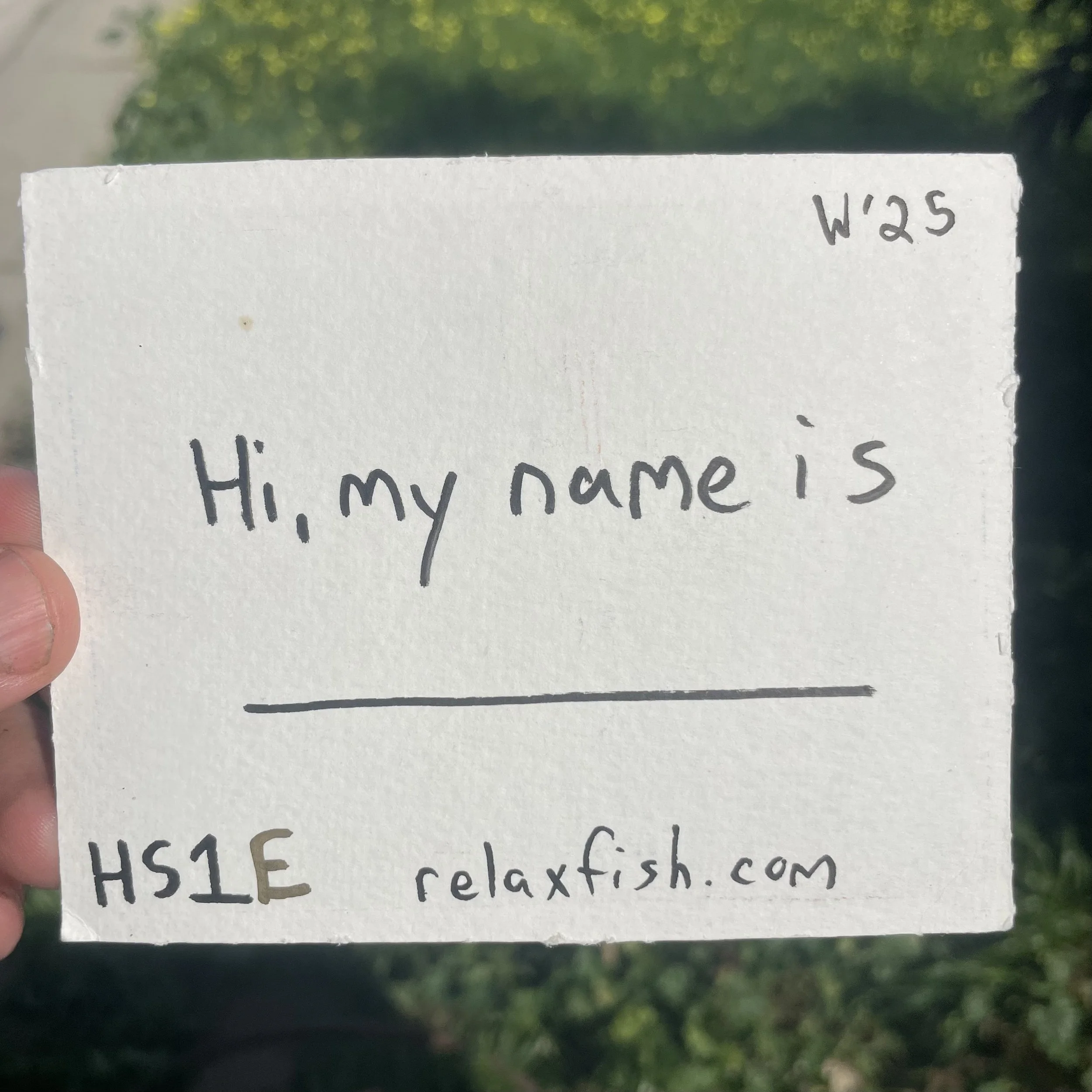 A person holding a white card outdoors with handwritten text that reads, 'Hi, my name is' and a blank line, with a website address relaxfish.com, and a code W25 in the top right corner.