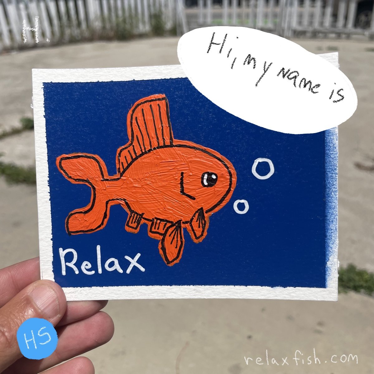 An illustrated orange fish with fins and a tail, drawn on a piece of paper, saying "Relax" at the bottom. A speech bubble says, "Hi, my name is." The drawing is held outdoors with a sandy background and a white fence.