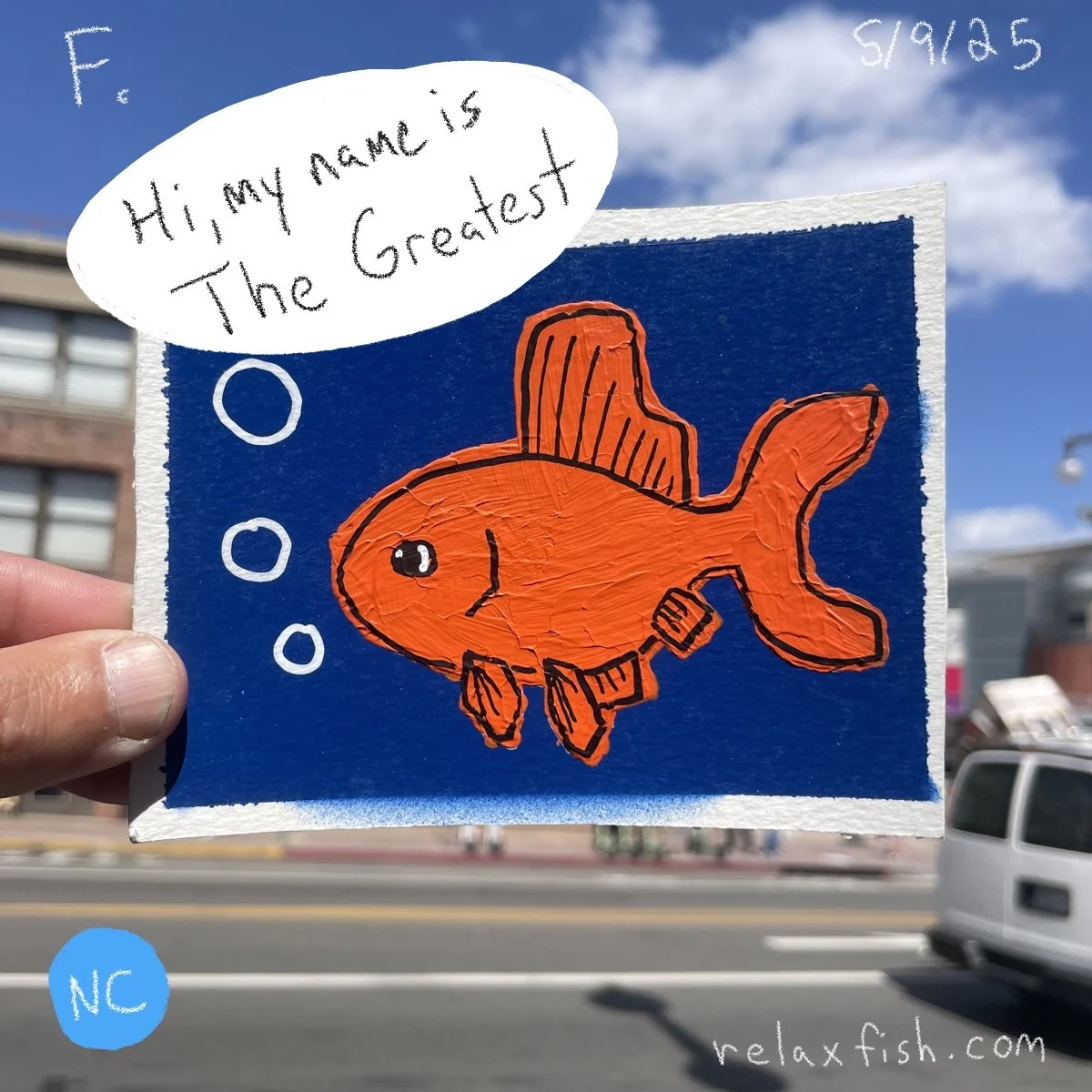 Hand holding a drawing of a goldfish with a speech bubble saying "Hi, my name is The Greatest." The drawing is on a blue background with a website link relaxfish.com written on it, and the image is outdoors with a street and buildings in the backgrou