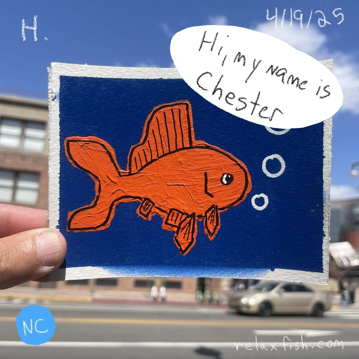 Colorful drawing of an orange fish with large fins on a navy blue background, with a speech bubble saying "Hi, my name is Chester" and bubbles around it. Hand holding the drawing outdoors with buildings and street in the background.