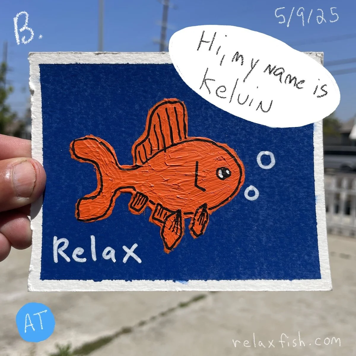Handheld drawing of an orange fish with black outlines on a blue background. Text in the image includes a speech bubble stating "Hi, my name is Kelvin," the word "Relax" at the bottom, and the date "5/9/23.5" in the top right corner. The drawing is o