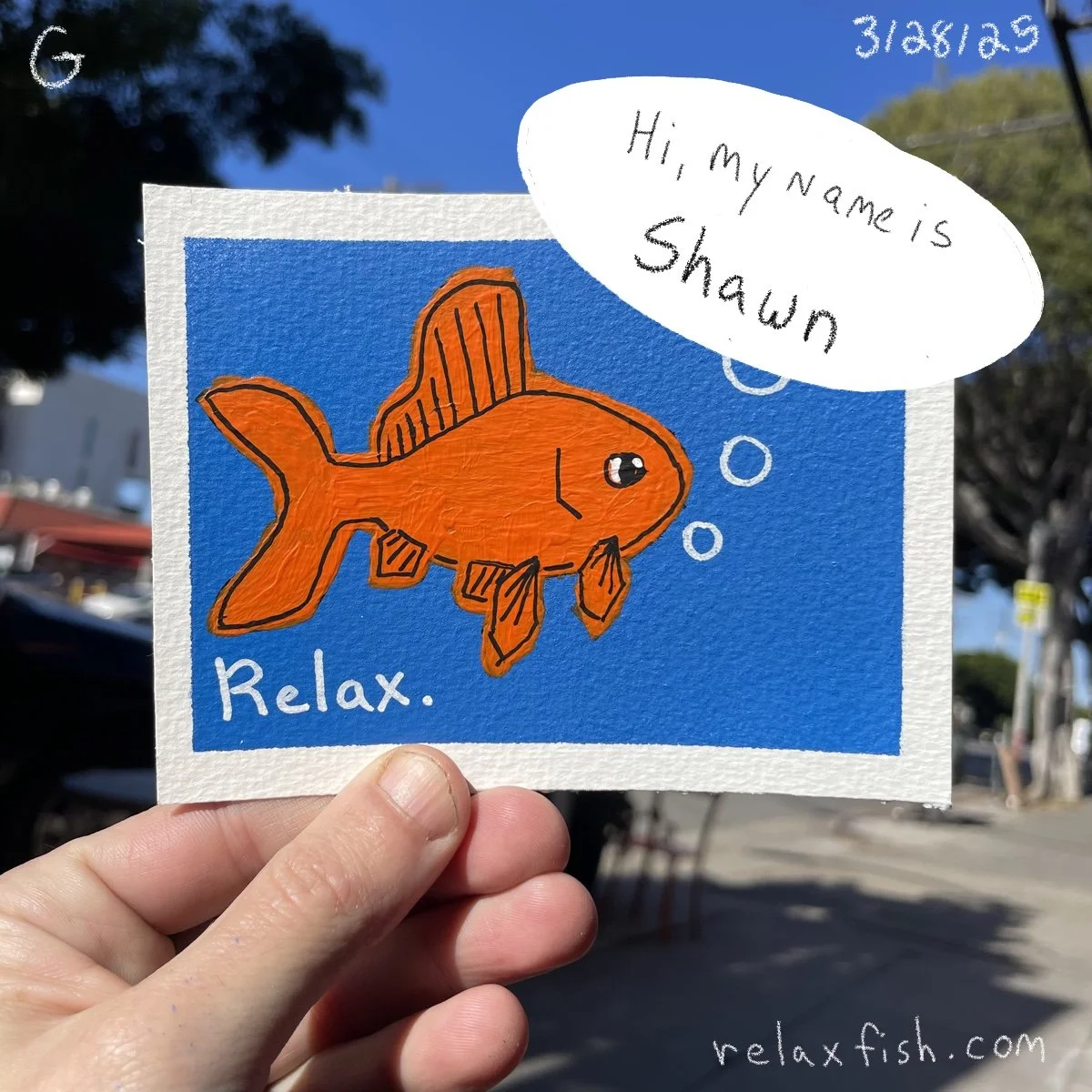 Hand holding a colorful drawing of an orange fish with the words "Relax." written on it, and a speech bubble saying "Hi, my name is Shawn." The drawing is outdoors with trees and buildings in the background.