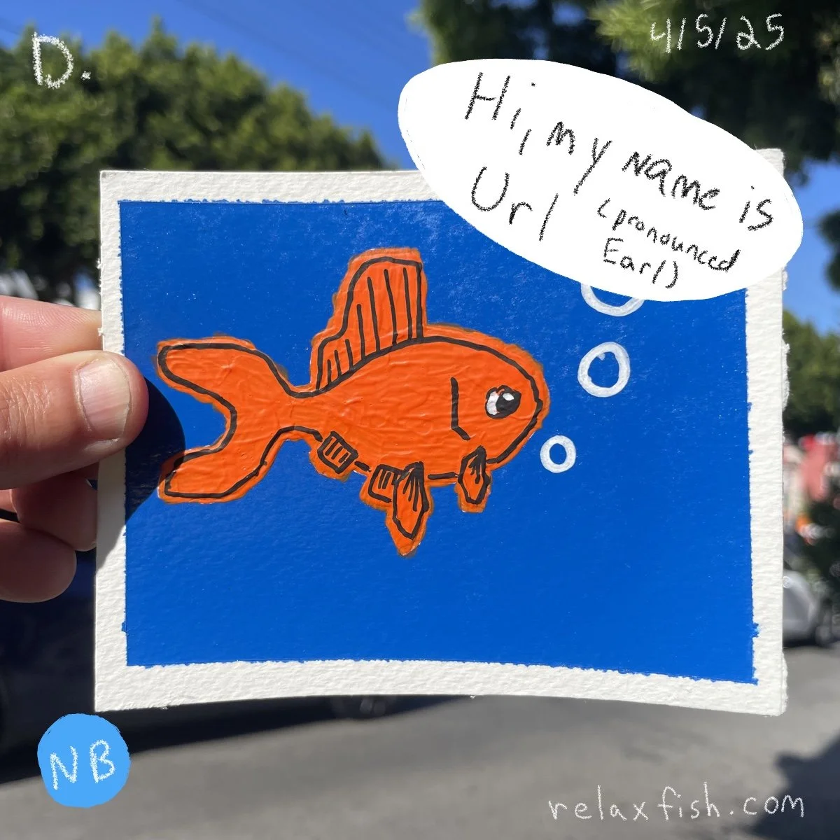 Hand holding a drawing of an orange goldfish with fins and a tail against a blue background. A speech bubble states, "My name is Ur! (pronounced Early)." The image features a white border with a small blue circle labeled "NB" in the bottom left corne