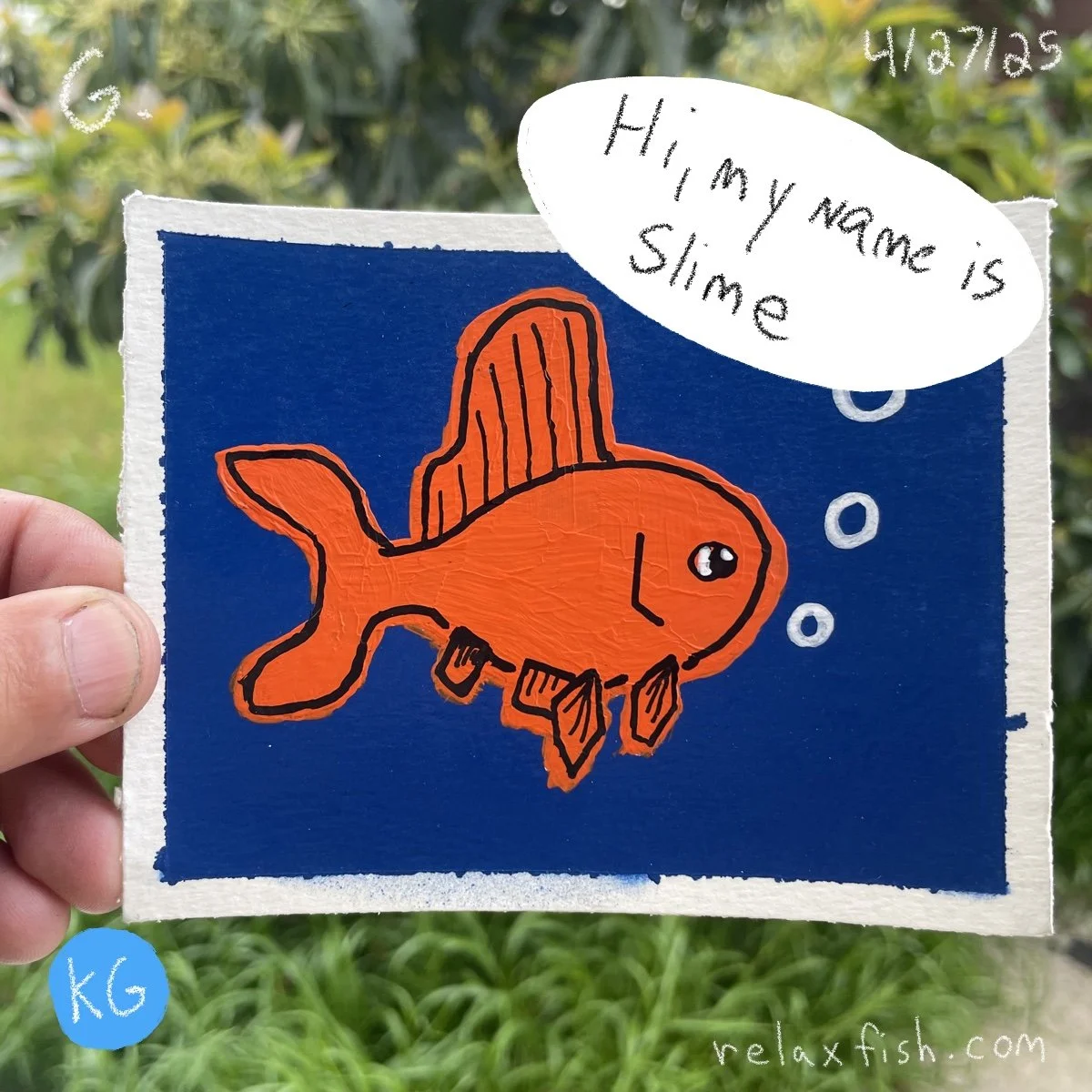 A hand holding a drawing of a goldfish with a blue background. The fish is orange with black outlines and a hanging cleo. There is a speech bubble saying, 'Hi, my name is Slime'. The background shows greenery. There are some small white bubbles near 