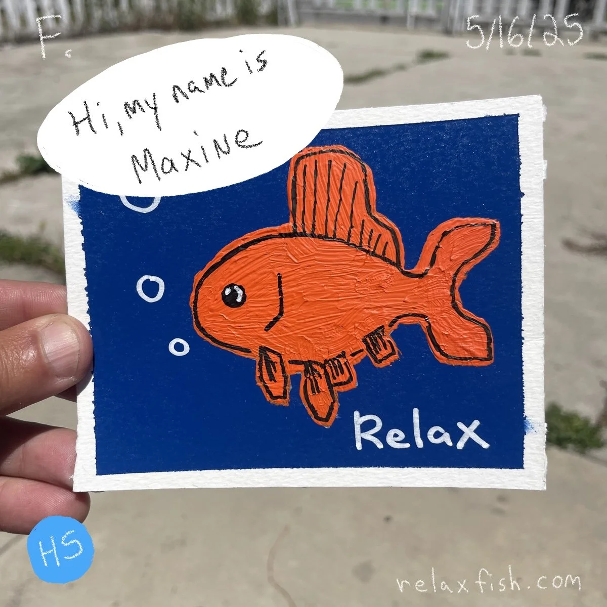 Drawing of an orange fish with black outlines and fins, floating underwater with bubbles, on a blue background. The fish is wearing a brown hat. Text on the drawing says "Relax" in white and a speech bubble says "Hi, my name is Maxine." Hand holding 
