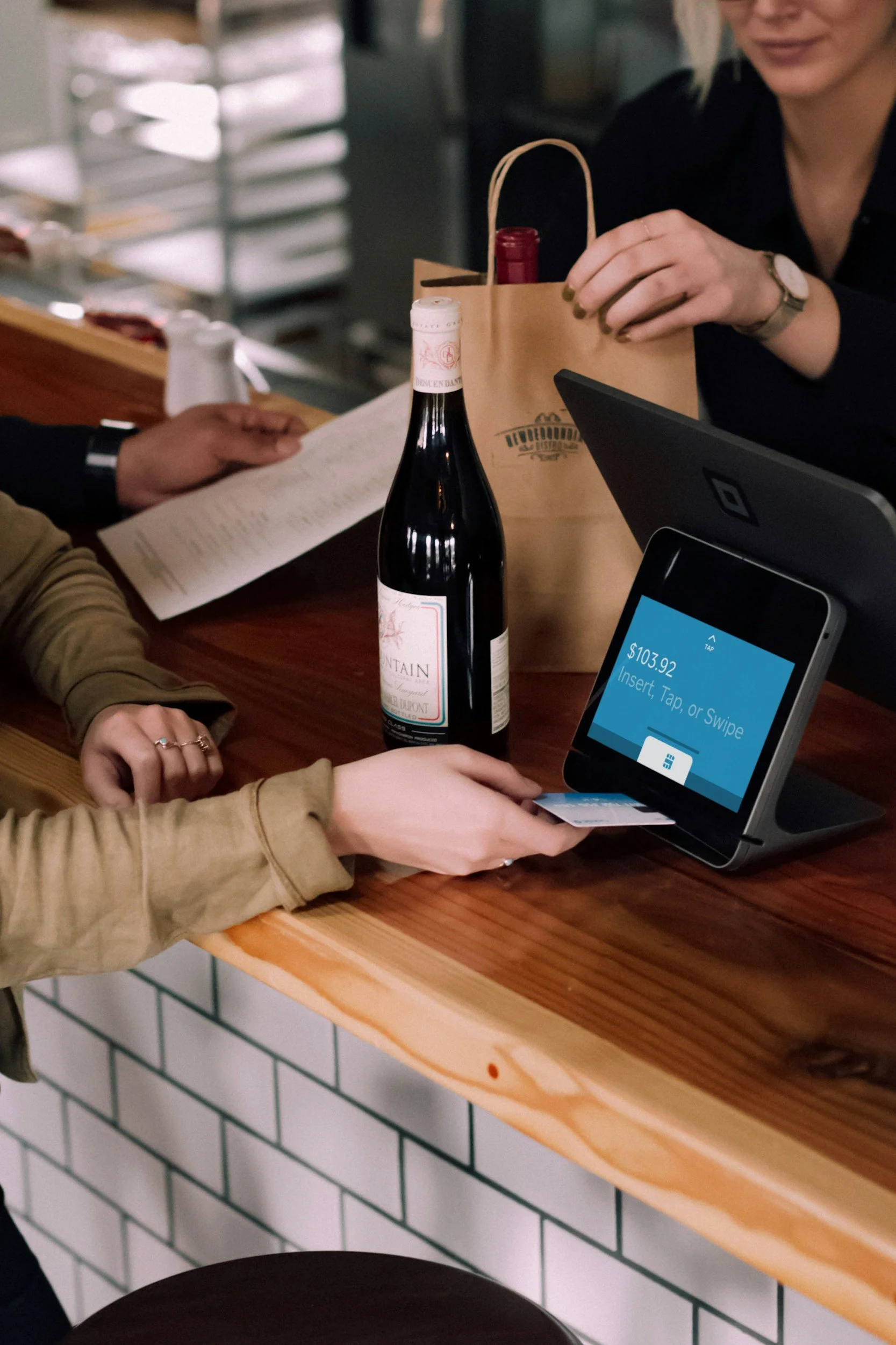 People at a wooden counter in a restaurant, with a bottle of wine, a small paper bag, a smartphone, and a payment terminal. One person is holding a credit card while another is making a payment via a tablet.