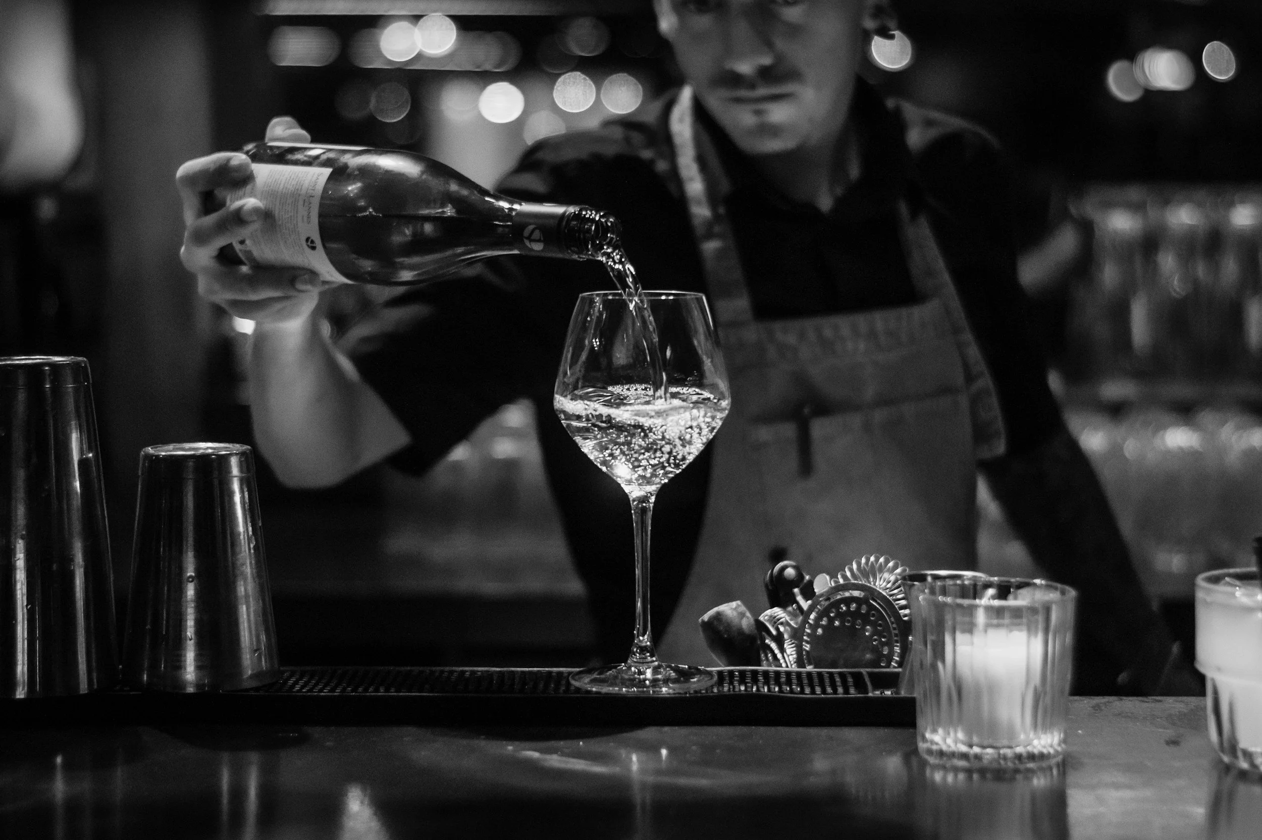 A bartender in a dark setting pours a beverage into a wine glass, with various bar tools and glasses on the bar counter.