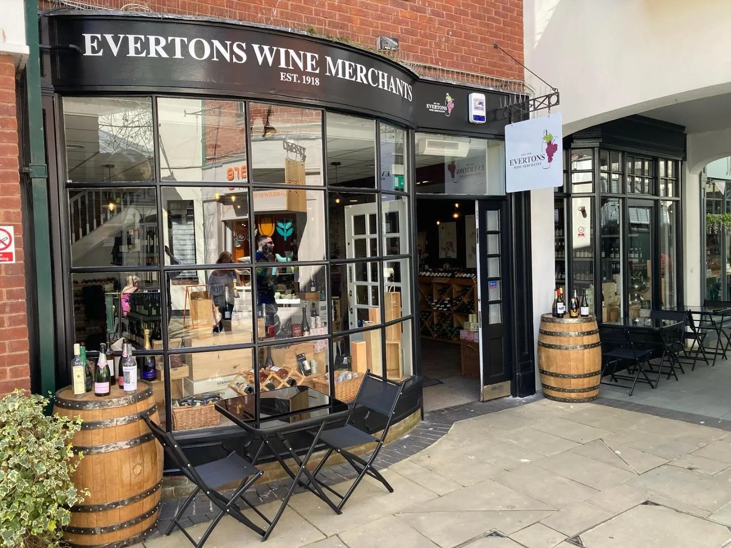 Exterior of Evertons Wine Merchants store with large glass windows, wine bottles displayed on barrels outside, and outdoor seating with black tables and chairs.