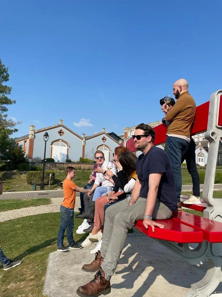 A group of people sitting on a red and gray structure in a park with a young boy talking to a woman. Two men are standing, drinking wine, and talking. The background shows a blue sky, a building, and some trees.
