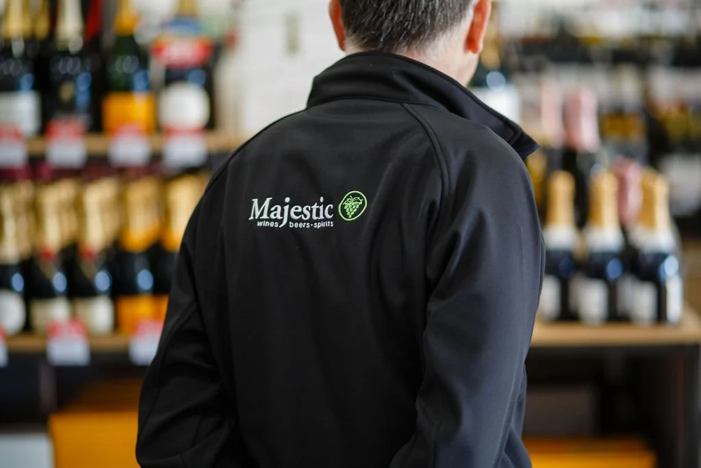 Person wearing a black jacket with a Majestic Wines logo, browsing alcohol bottles in a store.