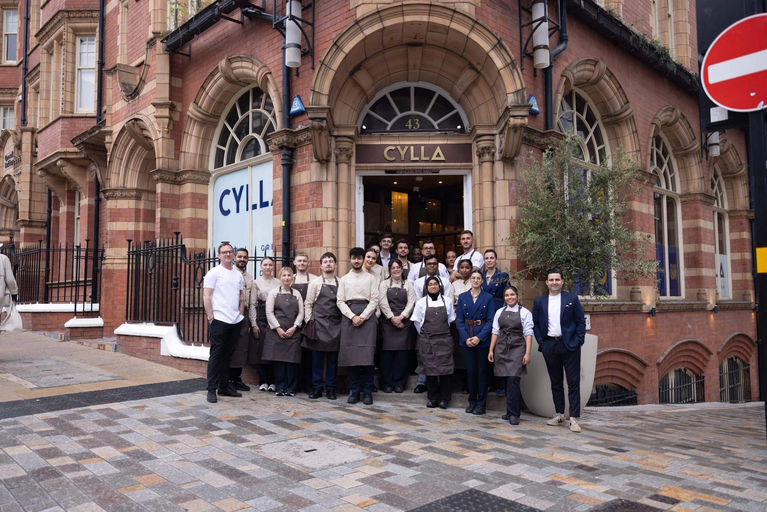 A group of restaurant staff and chefs standing outside a red brick building labeled CYLLA, posing for a photo. The building has large arched windows and a sign with the street number 43 above the entrance.