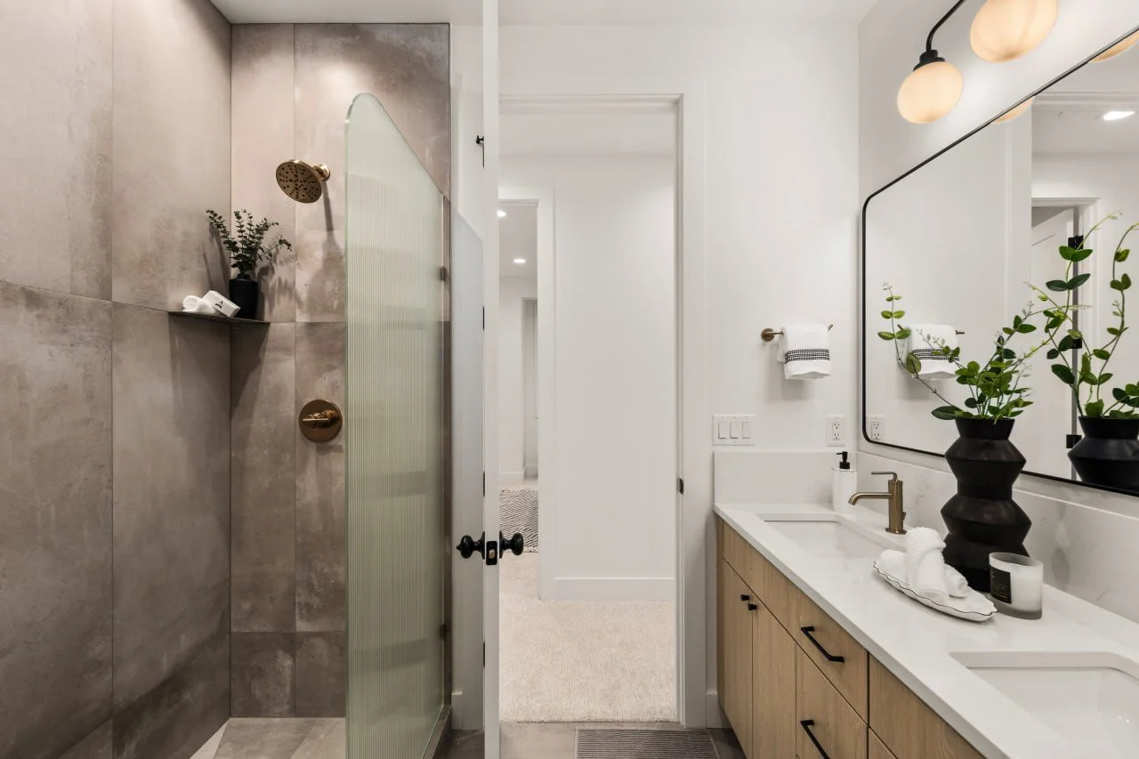 Modern bathroom with a shower area featuring large gray tiles and a brass shower head, a white vanity with black handles, a large mirror, and two black vases with green plants.