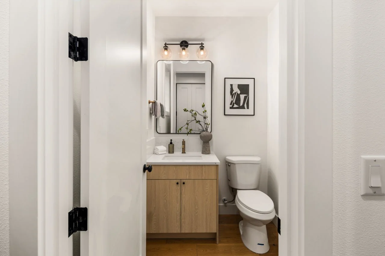 Small bathroom with white walls, wooden vanity with a mirror, small plants, and black and white artwork.