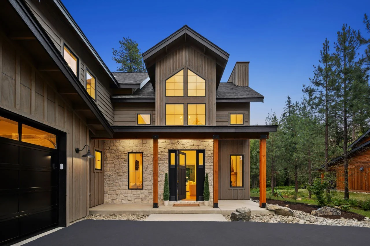Modern two-story house with large windows, wooden exterior, stone accents, and a black front door, surrounded by trees.