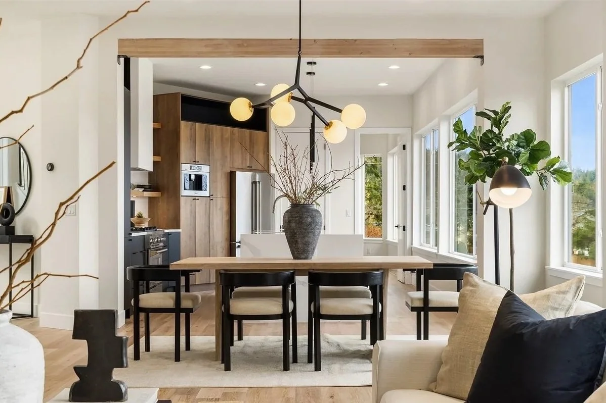 Open-plan living and dining area with large windows, a wooden dining table with six chairs, a vase with branches, modern lighting fixtures, and a glimpse of the kitchen with wood cabinets and stainless steel appliances.
