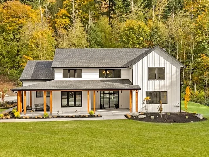 Modern two-story house with white vertical siding, black window frames, a covered porch with wooden support beams, and surrounded by a well-maintained lawn and trees with colorful fall foliage.