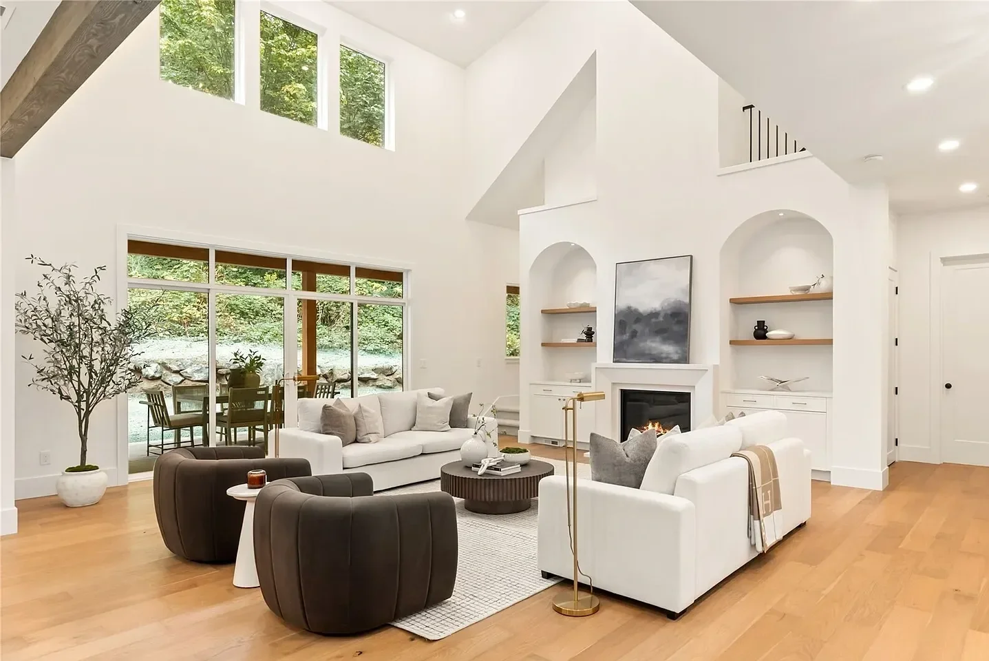 Living room with high vaulted ceiling, large windows showing greenery outside, and hardwood floors. Features include white sofas, dark armchairs, a round coffee table, a fireplace, and built-in shelves with decorations.