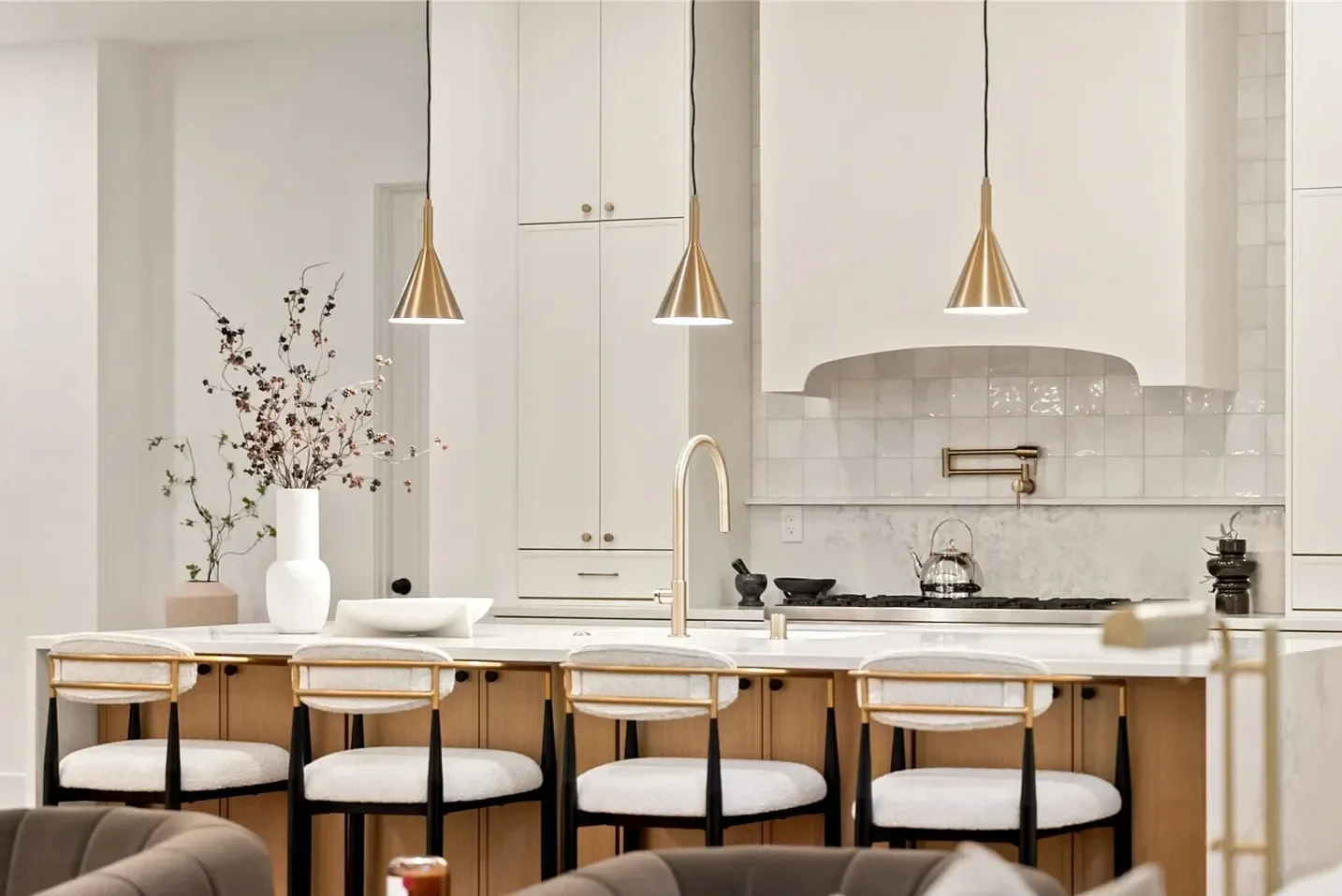 Modern kitchen with white cabinets, beige countertops, and a marble backsplash. Three gold pendant lights hang above a breakfast bar with four white upholstered chairs. Decor includes a white vase with branches, a teapot on the stove, and a black mor