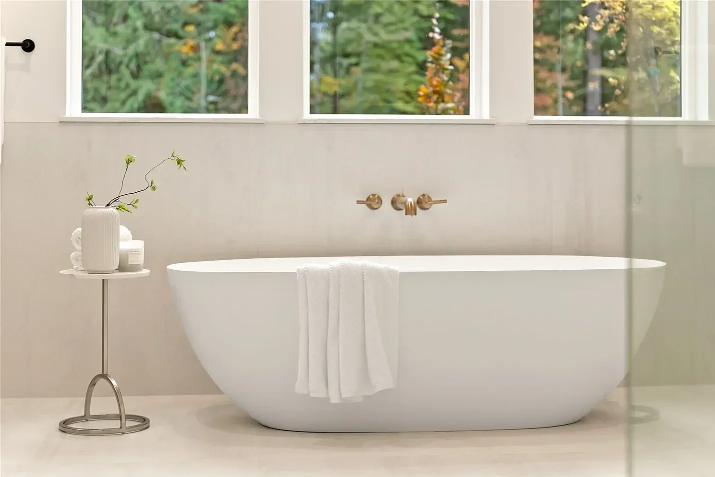 Modern white freestanding bathtub in a bright bathroom, with a towel hanging on it, a side table with a vase and skincare products, and large windows showing trees outside.