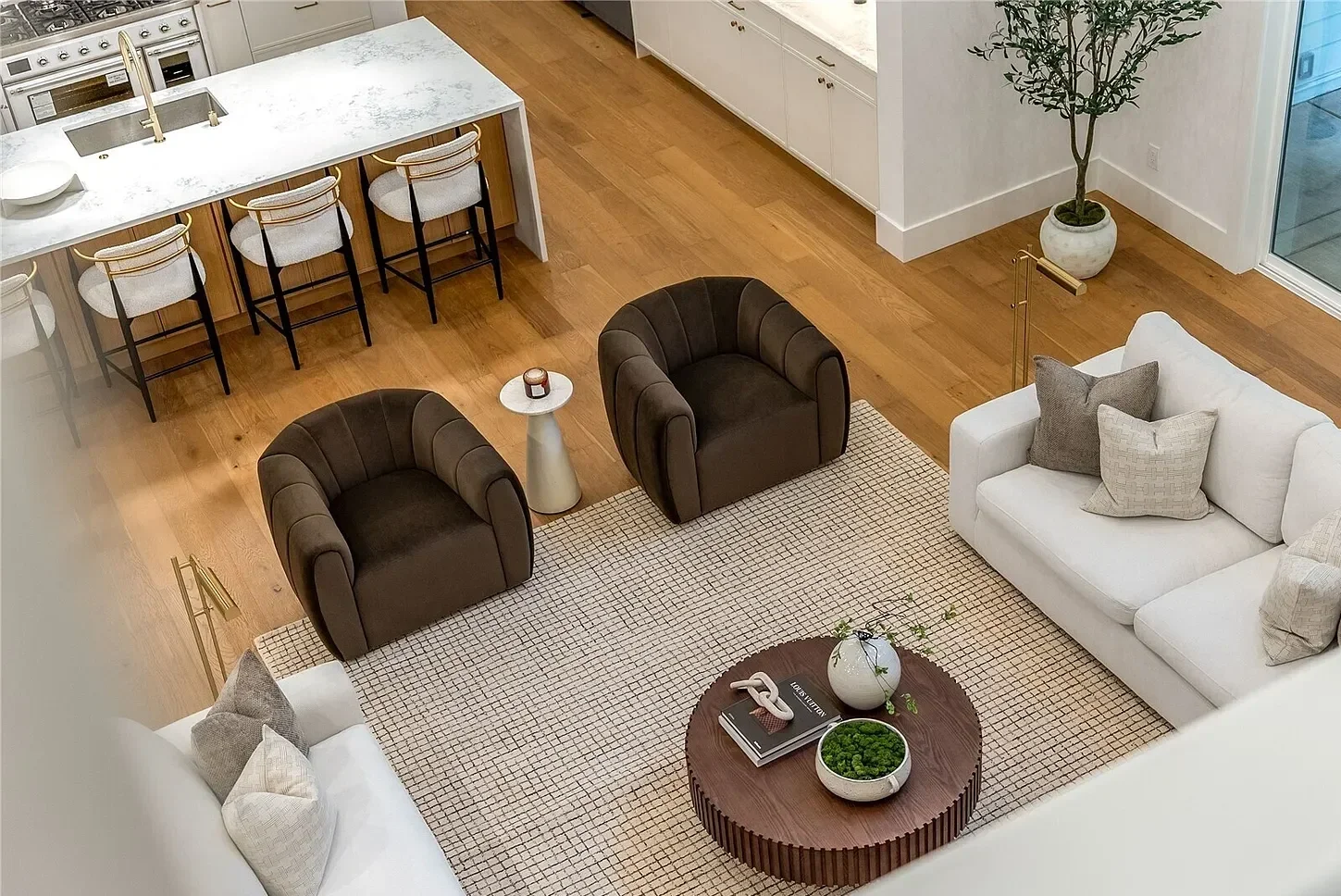 A modern living room with a white sofa, three beige and black armchairs, a round wooden coffee table with decorative items, and a large potted plant near a glass door. The area includes a checkered rug and a side table with a candle.