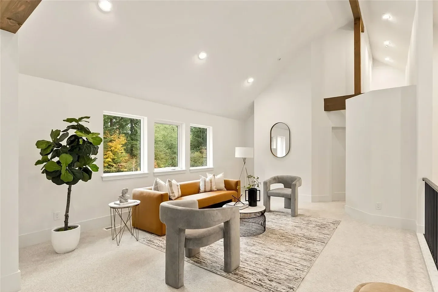 Living room with beige carpet, modern furniture, large windows showing trees outside, white walls, and ceiling lights.