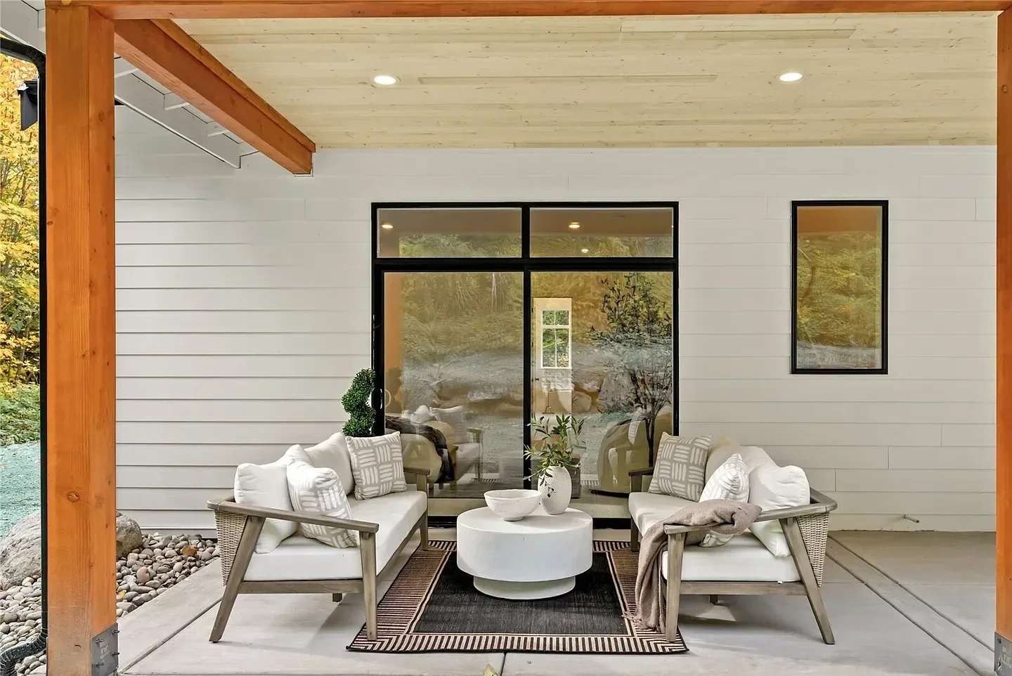 Covered outdoor patio with two white sofas, a round white coffee table, decorative pillows, and potted plants, with a glass sliding door and window on a white wall.