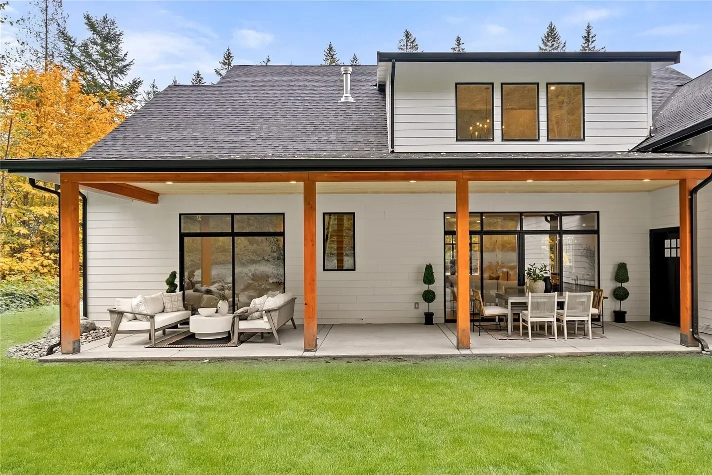 Modern two-story home with large covered patio, outdoor furniture, and a green lawn, surrounded by trees.