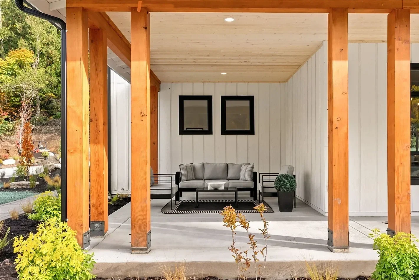 Outdoor sitting area with a gray sofa, two black chairs, a black coffee table, framed black and white artwork on a white wooden wall, potted plant, and a black and white striped rug, with a porch supported by wooden beams.