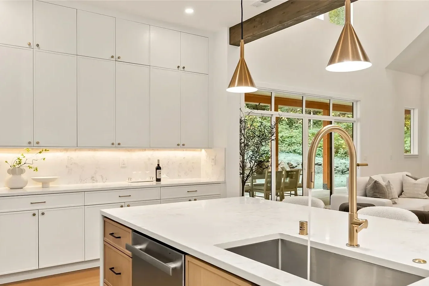Modern kitchen with white cabinets, a white marble backsplash, and a white marble countertop island with a gold faucet. Two pendent lights hang above the island. Large windows and a sliding glass door reveal an outdoor patio and greenery.