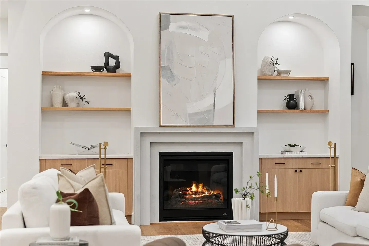 Living room with white walls and a central fireplace, surrounded by built-in shelves with decorative objects. A large abstract painting is hung above the fireplace, and the room features white sofas with pillows, a black coffee table with magazines, 